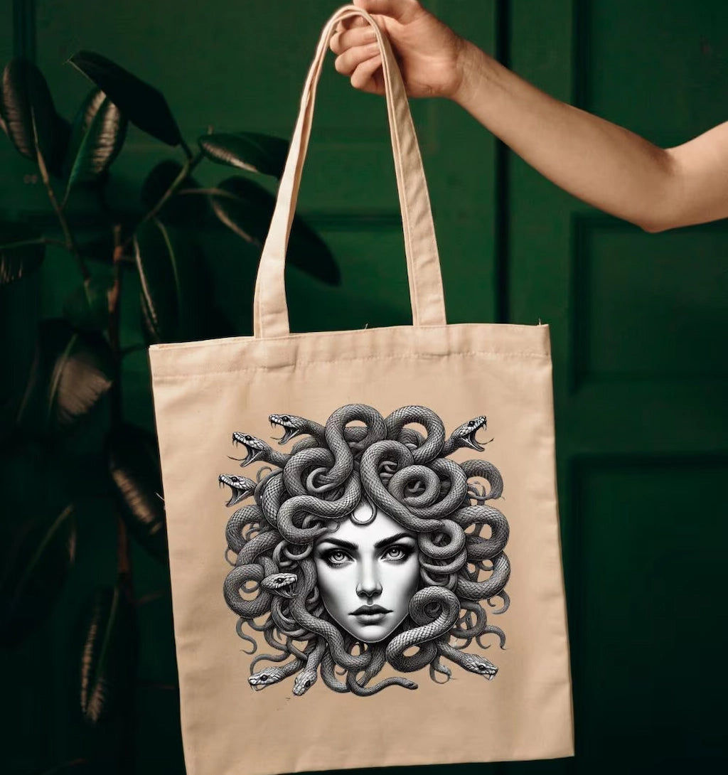 Beige tote bag with detailed Medusa artwork, featuring snake hair, held indoors by a hand