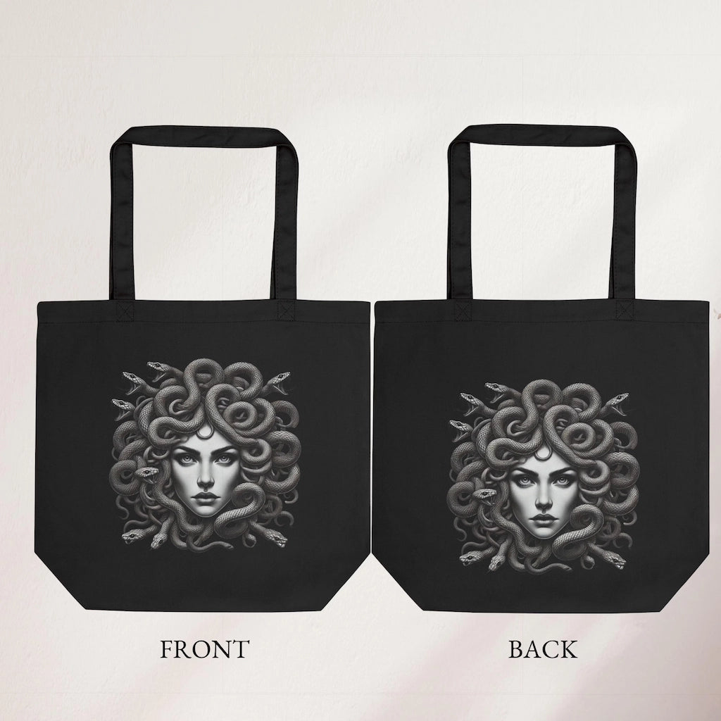 Black tote bag with detailed Medusa artwork, featuring snake hair, held indoors by a hand
