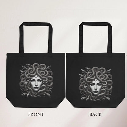 Black tote bag with detailed Medusa artwork, featuring snake hair, held indoors by a hand