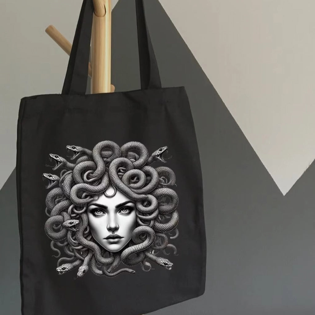 Black tote bag with detailed Medusa artwork, featuring snake hair, held indoors by a hand