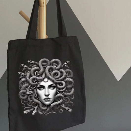 Black tote bag with detailed Medusa artwork, featuring snake hair, held indoors by a hand