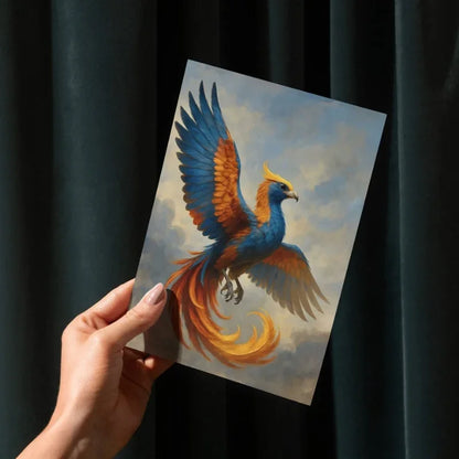 Hand holding fantasy art print of a vibrant blue and orange phoenix against a cloudy sky